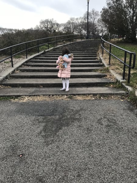 A girl wearing a pink puffer jacket with fur collar, white tights and flats is pictured at the bottom of a concrete stairwell in a park holding her toys, which obscure her face.
