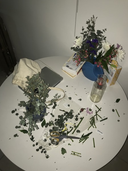 Newly-cut leaves and stems from a just-purchased bouquet cover a round white table next to full and empty vases, a box of straws, books, a tote, a laptop computer.