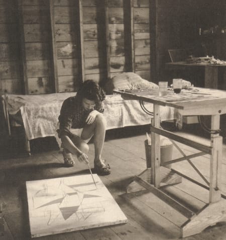 A sepia photo of a woman painting on the floor in a bare room with a table and bed
