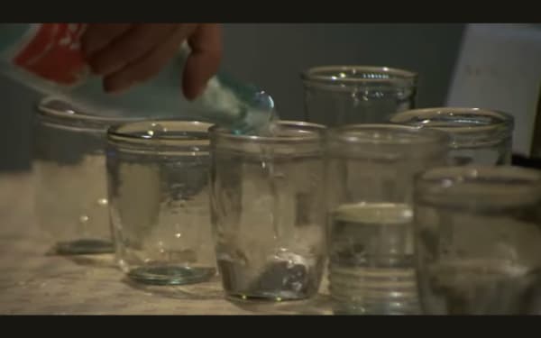 Screenshot from How Near How Far (1972) showing various jars and glasses and someone pouring a clear liquid into them from a bottle, probably vodka