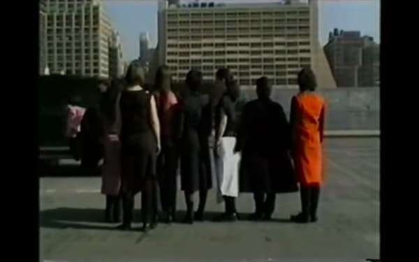 Screenshot from BLESS video showing a group of people facing away from camera wearing black with one person in red