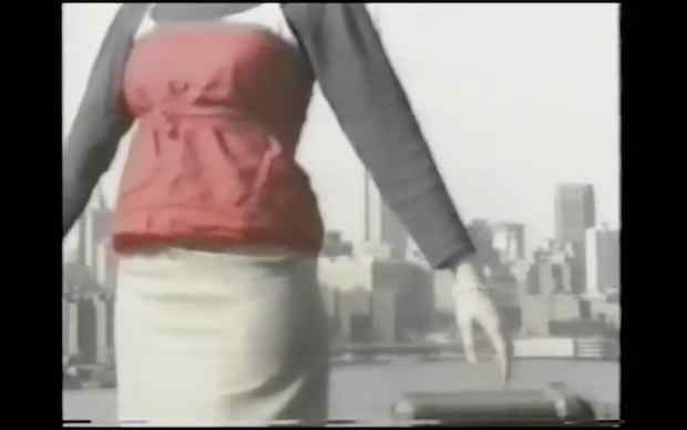 From Susan Cianciolo's Films and Collaborations 1996-2012, shot from neck to thighs, a woman in a long sleeve top layered under white camisole layered under red sleeveless top tied at the waist, in a beige skirt, stands in front of a cityscape that looks like NYC