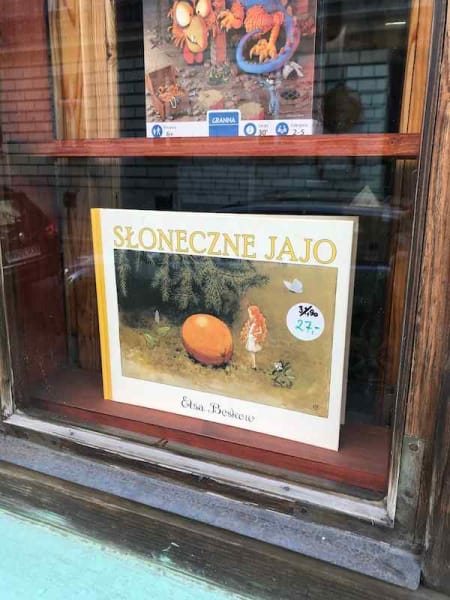 Photo of children's book in a window display titled Słoneczne Jajo, translates as The Sun Egg, showing a tiny girl with orange hair standing next to a lemon-sized orange fruit