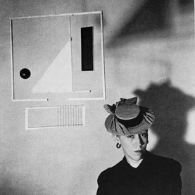 Black and white photo of a woman in a strange hat underneath a miniamlist geometric wall scultpure, xenia cage