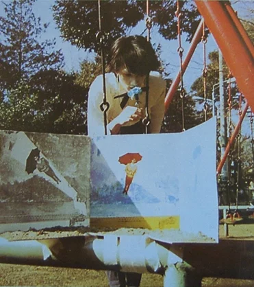 Photo of band Pale Cocoon, showing a woman maybe speaking into something in a playground behind what may be an open record