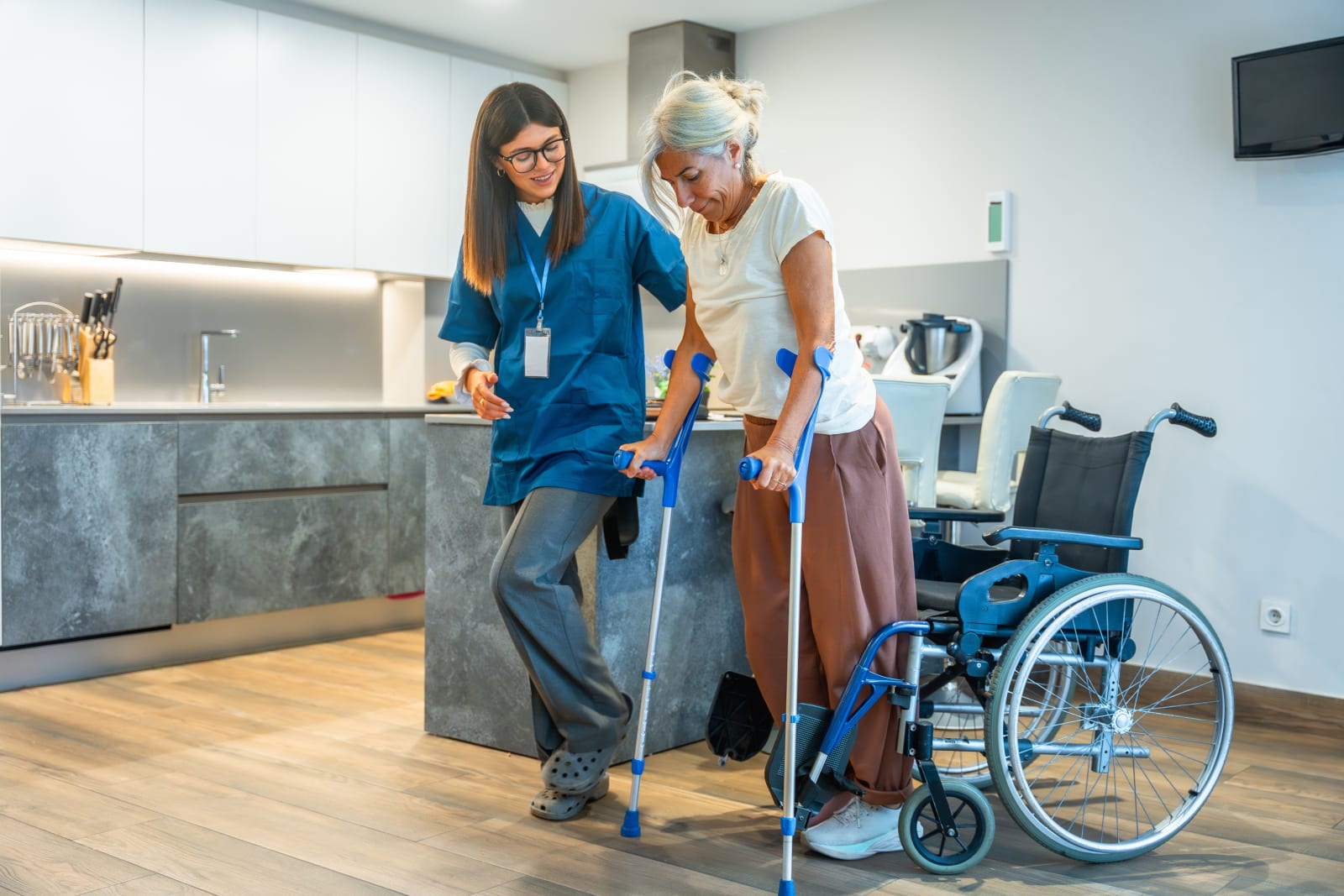 Caregiver helping an elderly woman walking with a wheeled walker through a sunlit kitchen