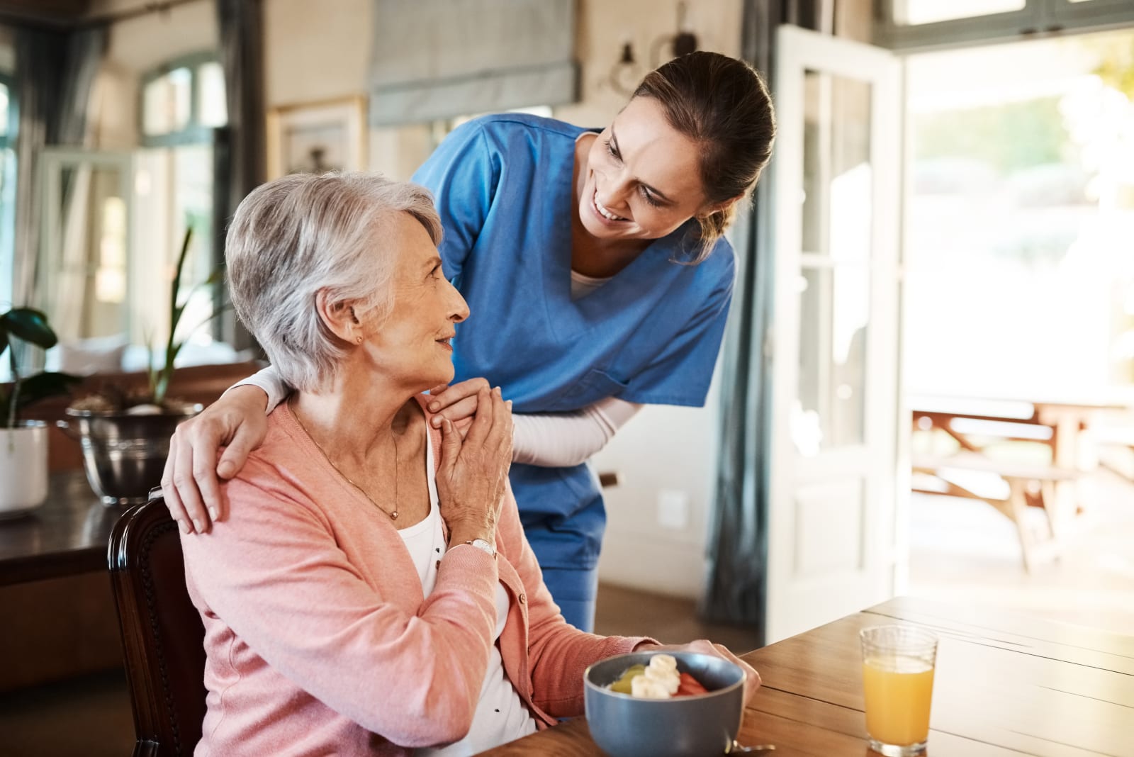 Caregiver resting a reassuring hand on an elderly woman's shoulder at the breakfast table