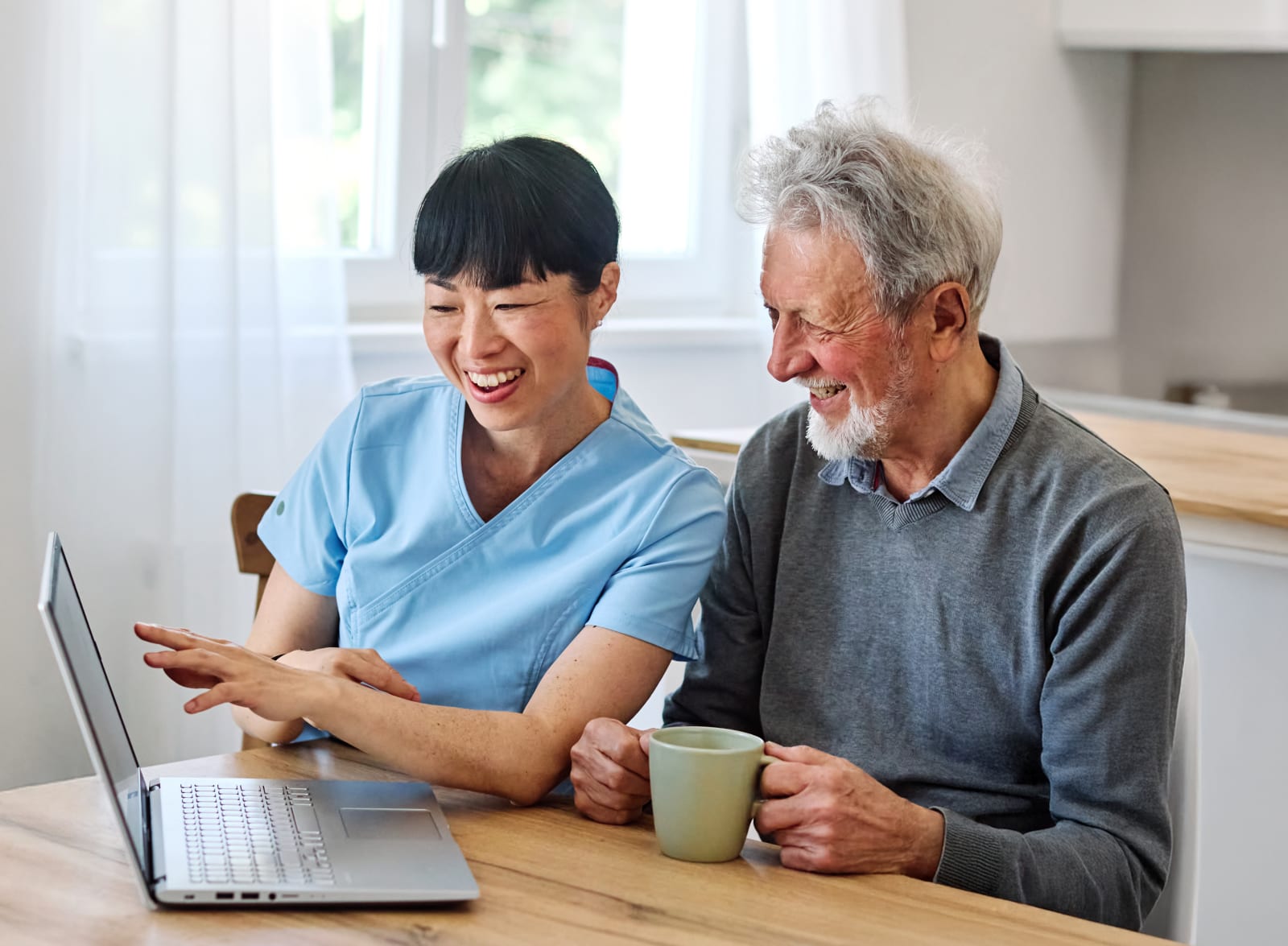 Caregiver and elderly man at a home table reviewing a laptop together over coffee