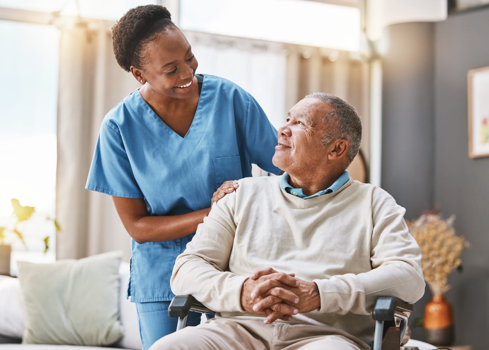 A caregiver sitting beside an elderly gentleman in a sunlit living room, both smiling at each other