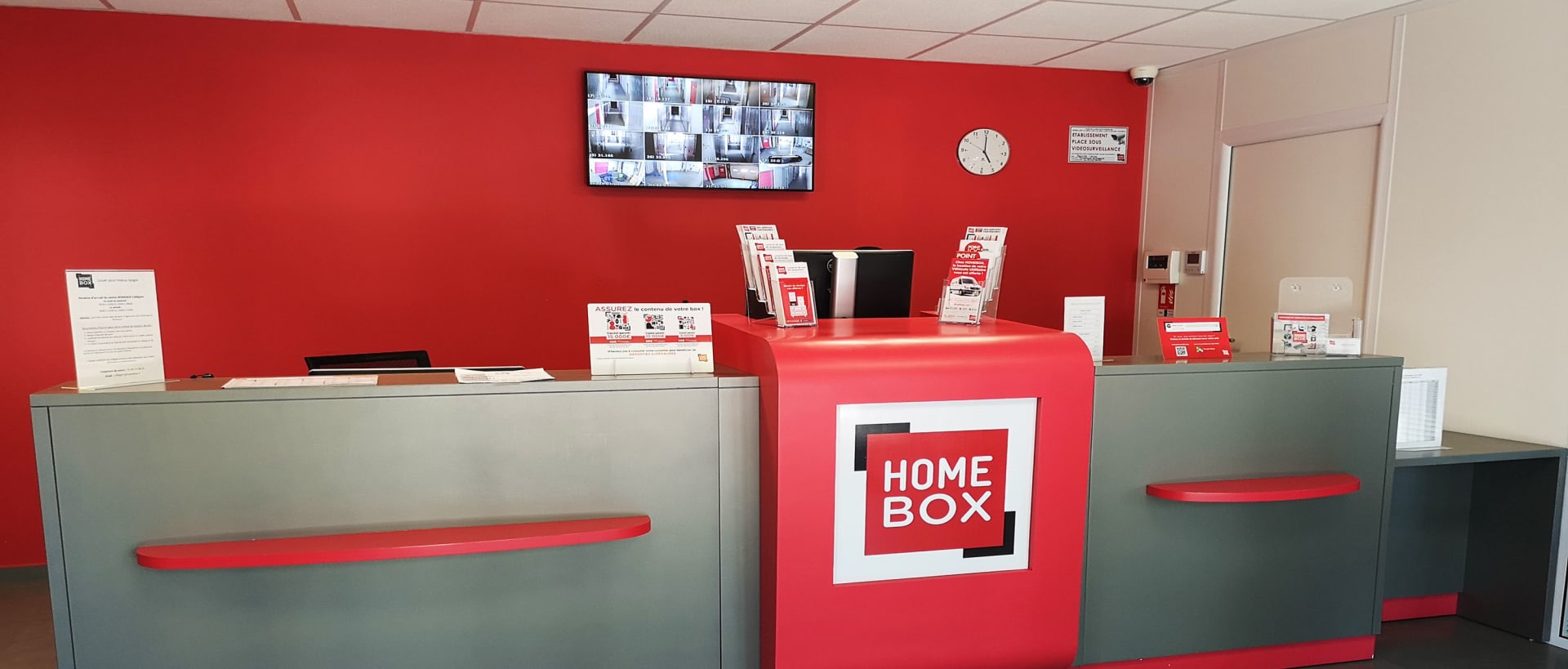 Comptoir d'accueil rouge et gris avec écran de vidéosurveillance et logo cube du centre HOMEBOX Vannes Nord