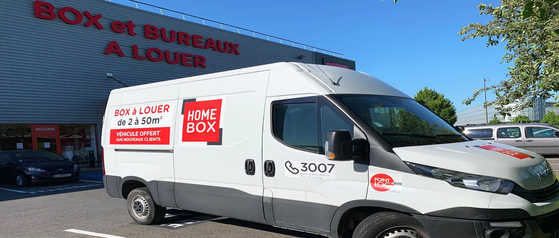 Camion utilitaire HOMEBOX devant la façade rouge "Box et bureaux à louer" du centre HOMEBOX Roissy, 383 rue de la Belle Etoile Parc des Nations à Roissy