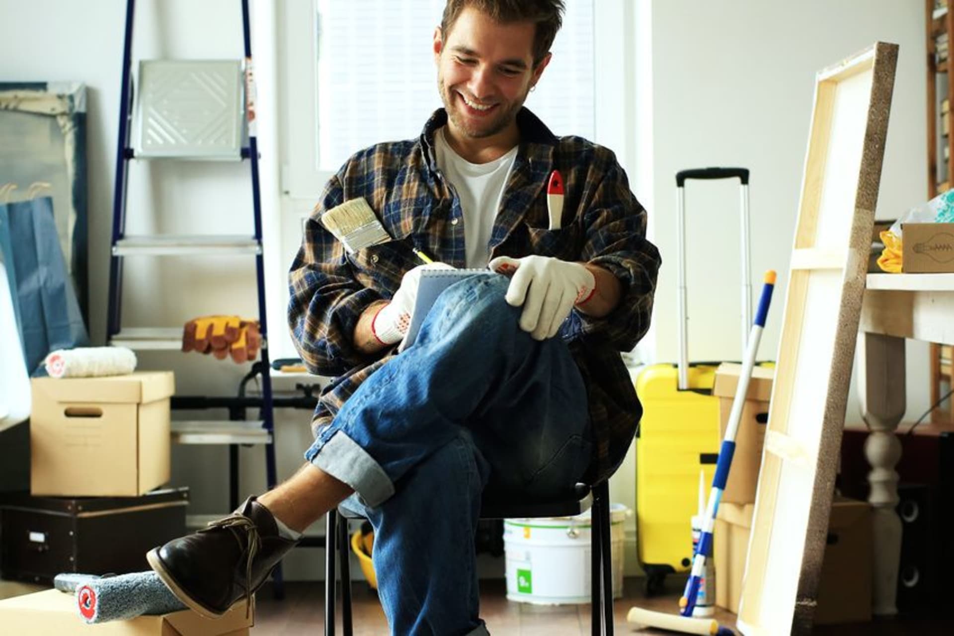 Homme souriant assis avec pinceau et calepin parmi des cartons et du matériel de nettoyage