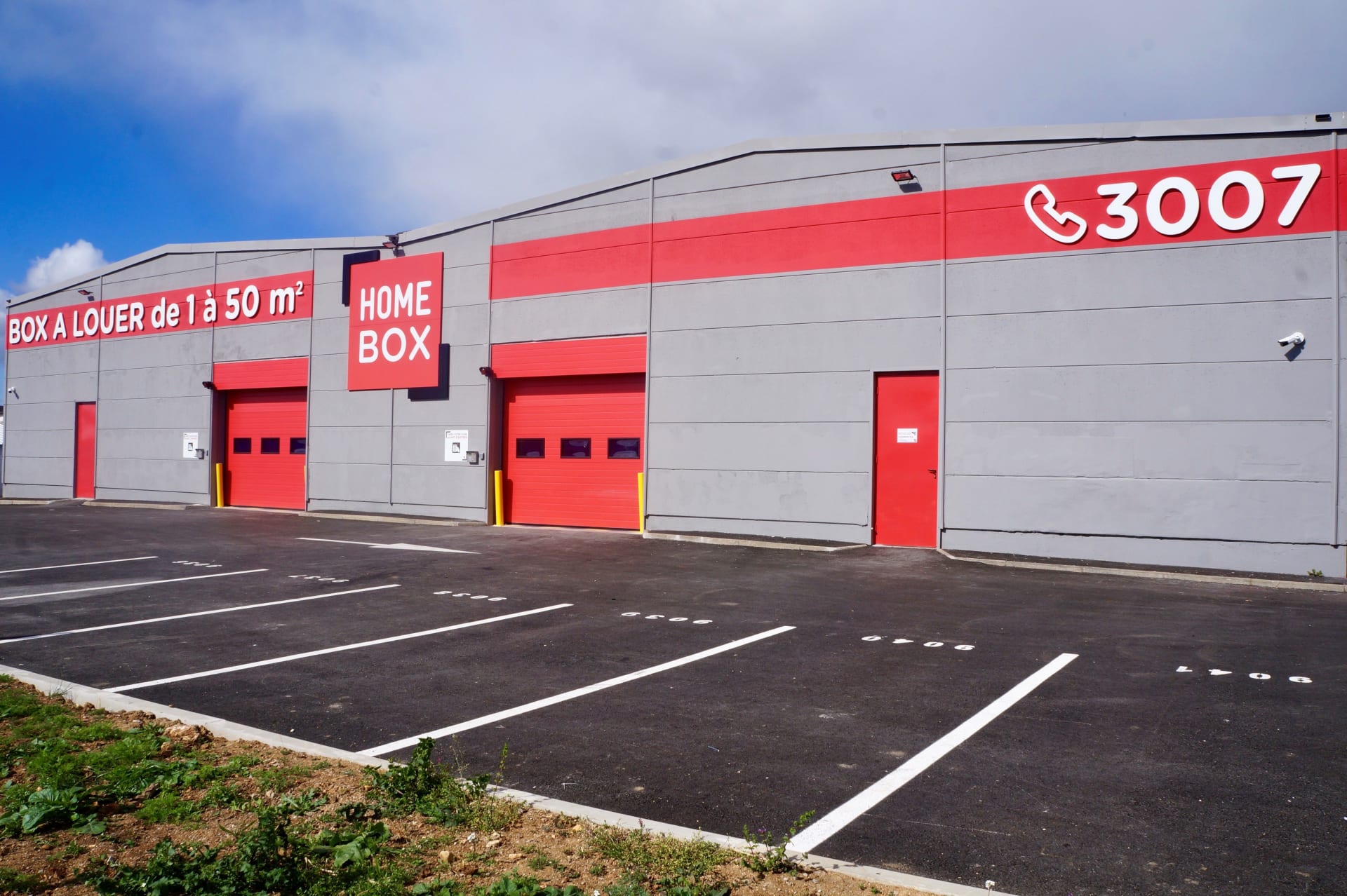 Façade grise avec portes de garage rouges et bandeau d'enseigne, HOMEBOX Montpellier, 925 Avenue du Marché Gare à Montpellier