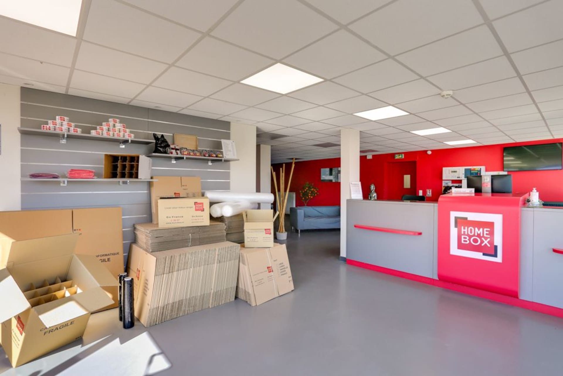 Accueil avec cartons de déménagement empilés, comptoir rouge et présentoirs de fournitures du centre HOMEBOX Rennes Donelière