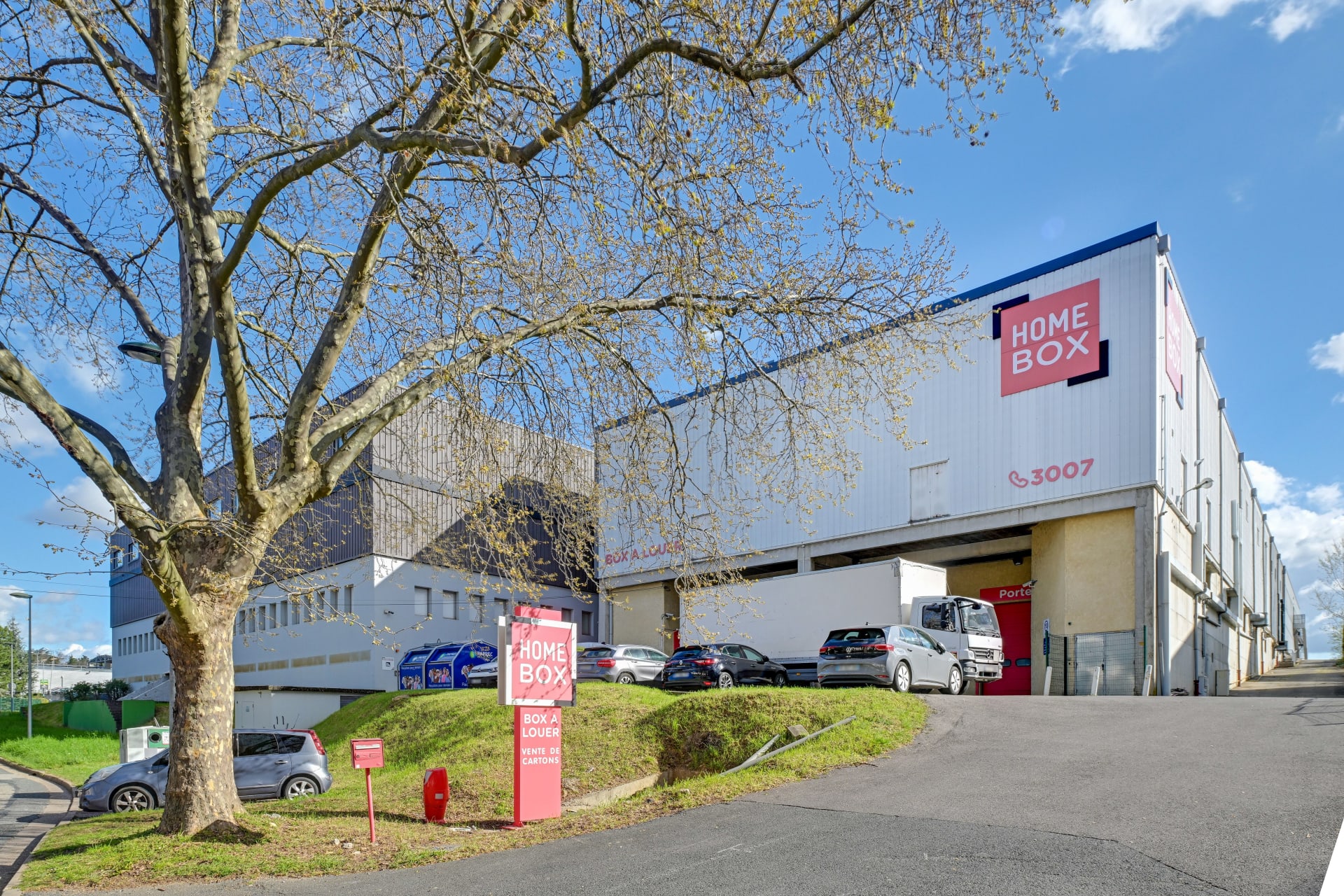 Vue extérieure du bâtiment avec enseigne HOMEBOX, parking et arbre au premier plan, HOMEBOX Lyon Ouest, 17 rue des Rosieristes à Champagne au Mont d'Or