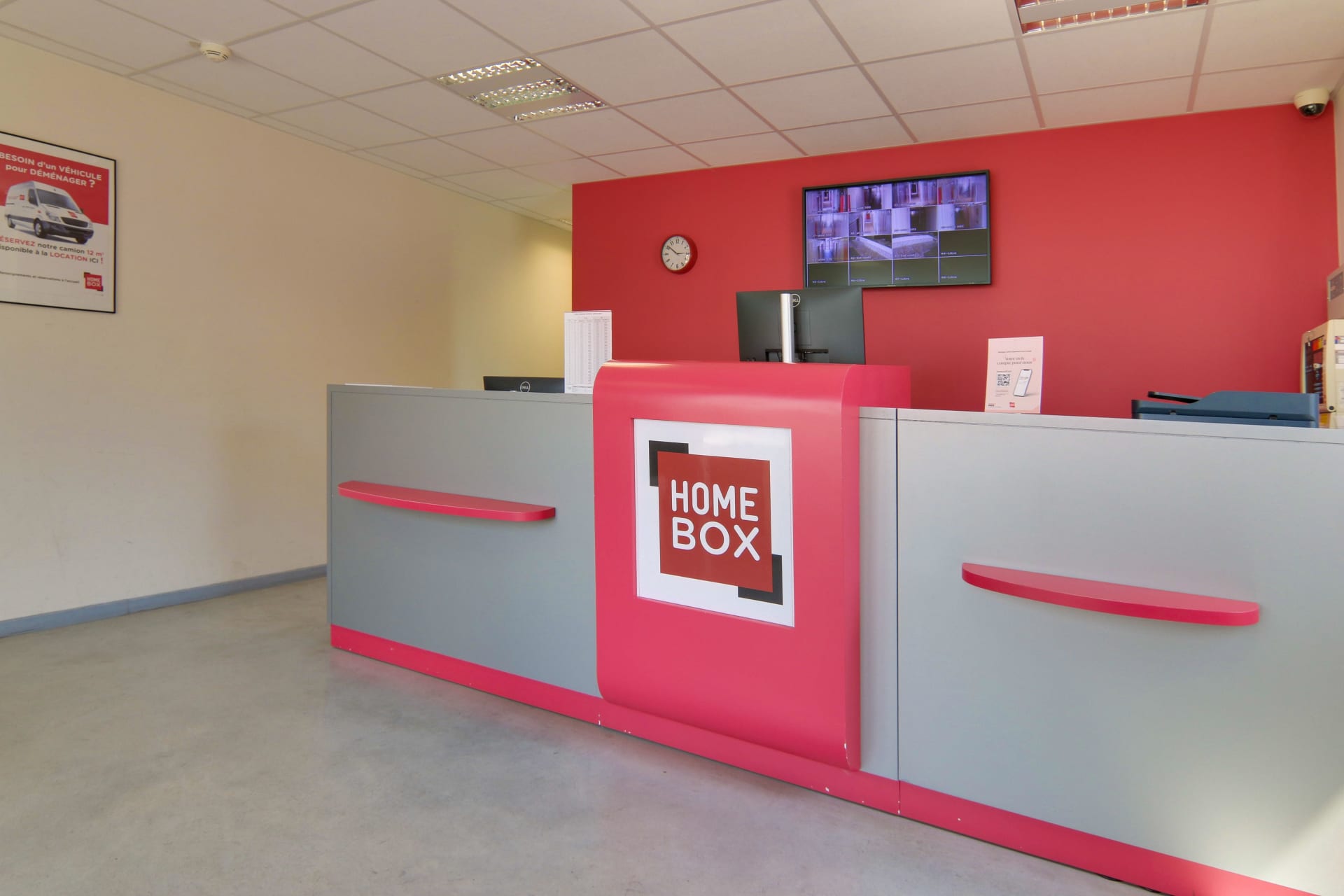 Comptoir d’accueil du centre HOMEBOX Avignon avec mur rouge et écran de vidéosurveillance.