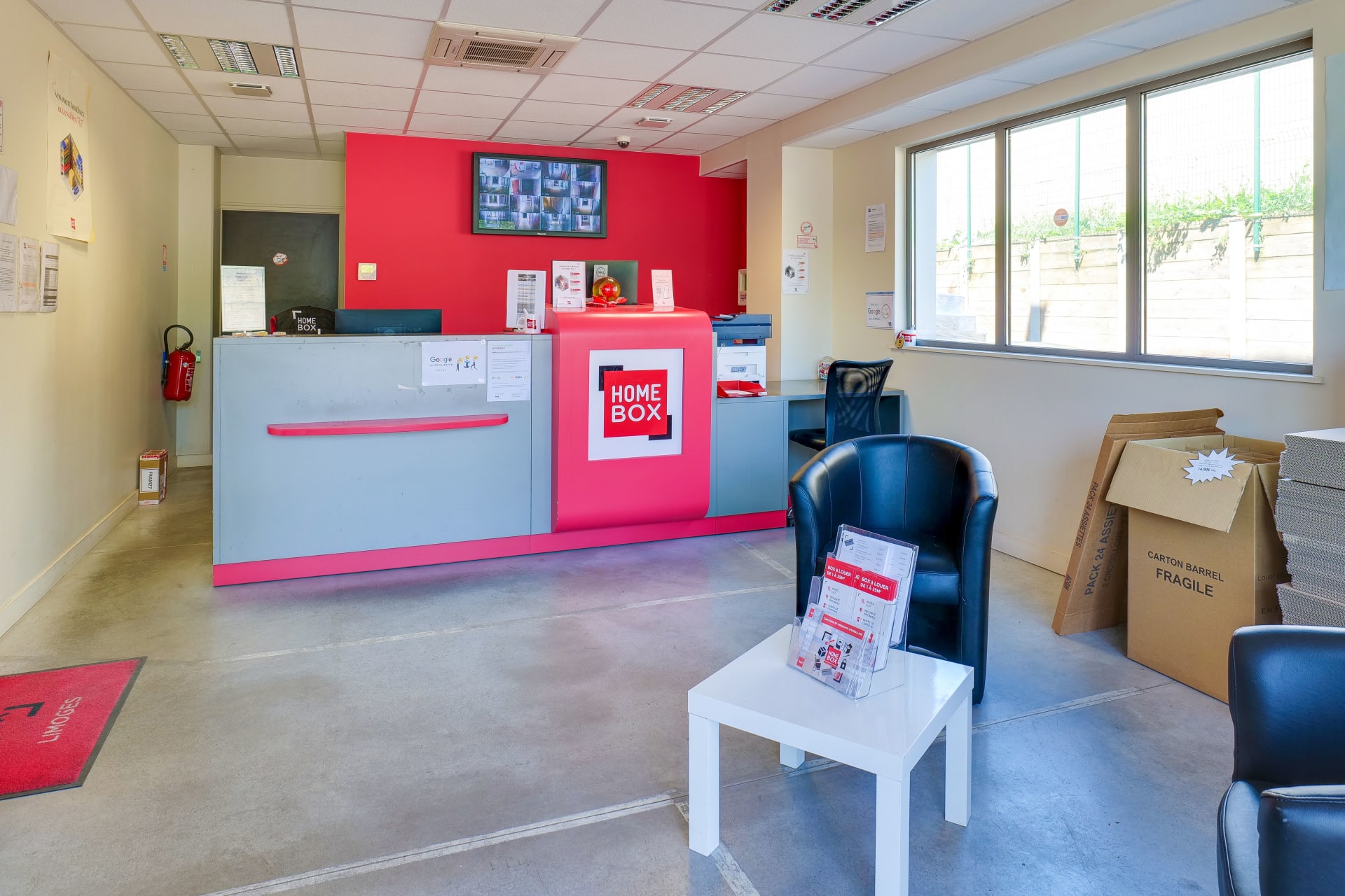 Bureau d'accueil avec comptoir rouge, fauteuils noirs et cartons d'emballage du centre HOMEBOX Limoges