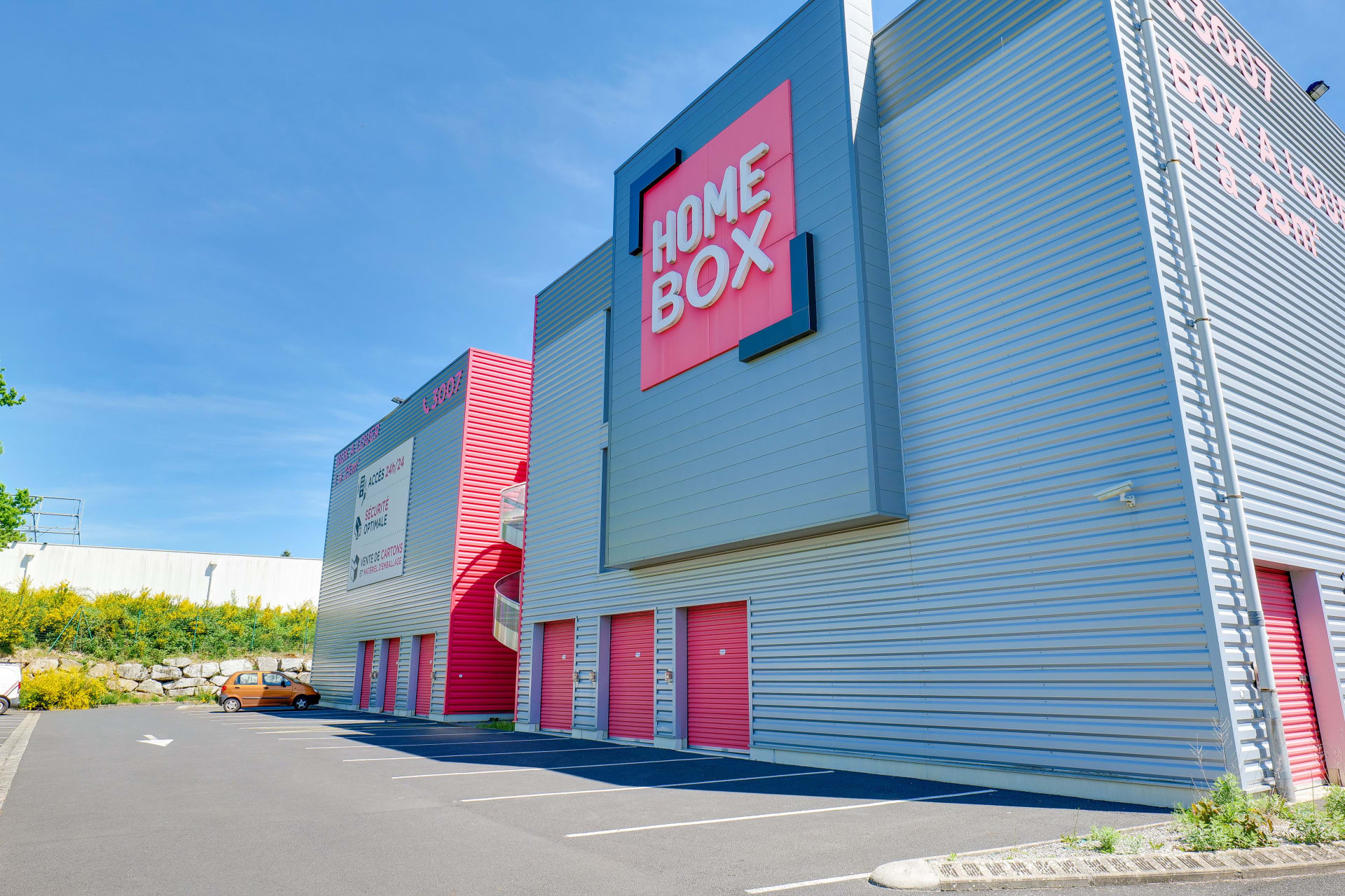 Façade grise avec enseigne HOMEBOX rose et rideaux de boxes rouges sous ciel bleu, HOMEBOX Limoges, 8 Allée Louis Daubenton ZAC Beaubreuil à Limoges