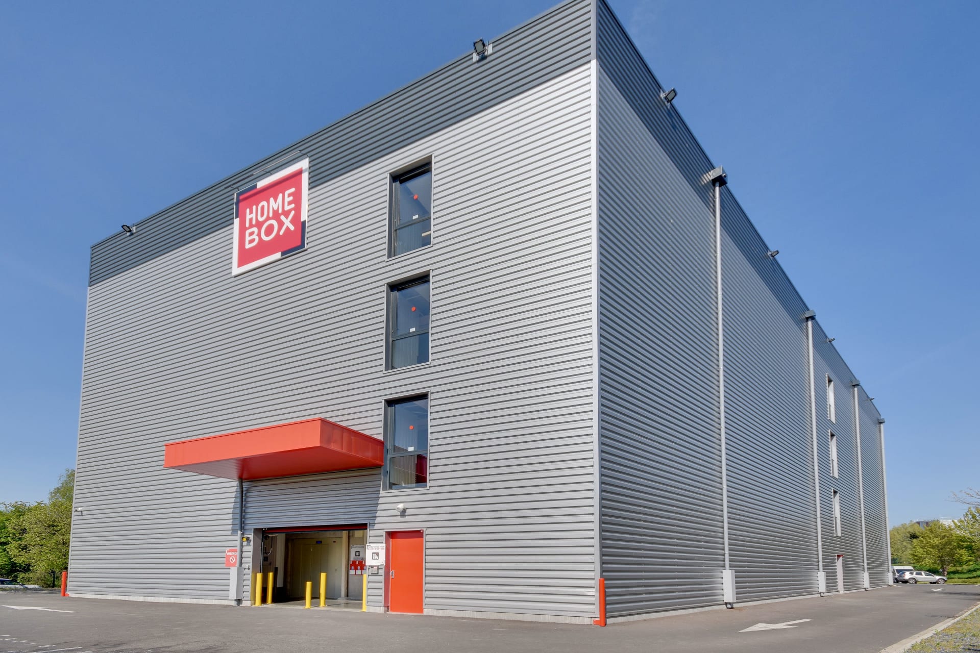 Façade d'angle bardage gris avec enseigne et auvent rouge sous ciel bleu, HOMEBOX Lille Lomme, Rue du Traité de Rome à Lomme