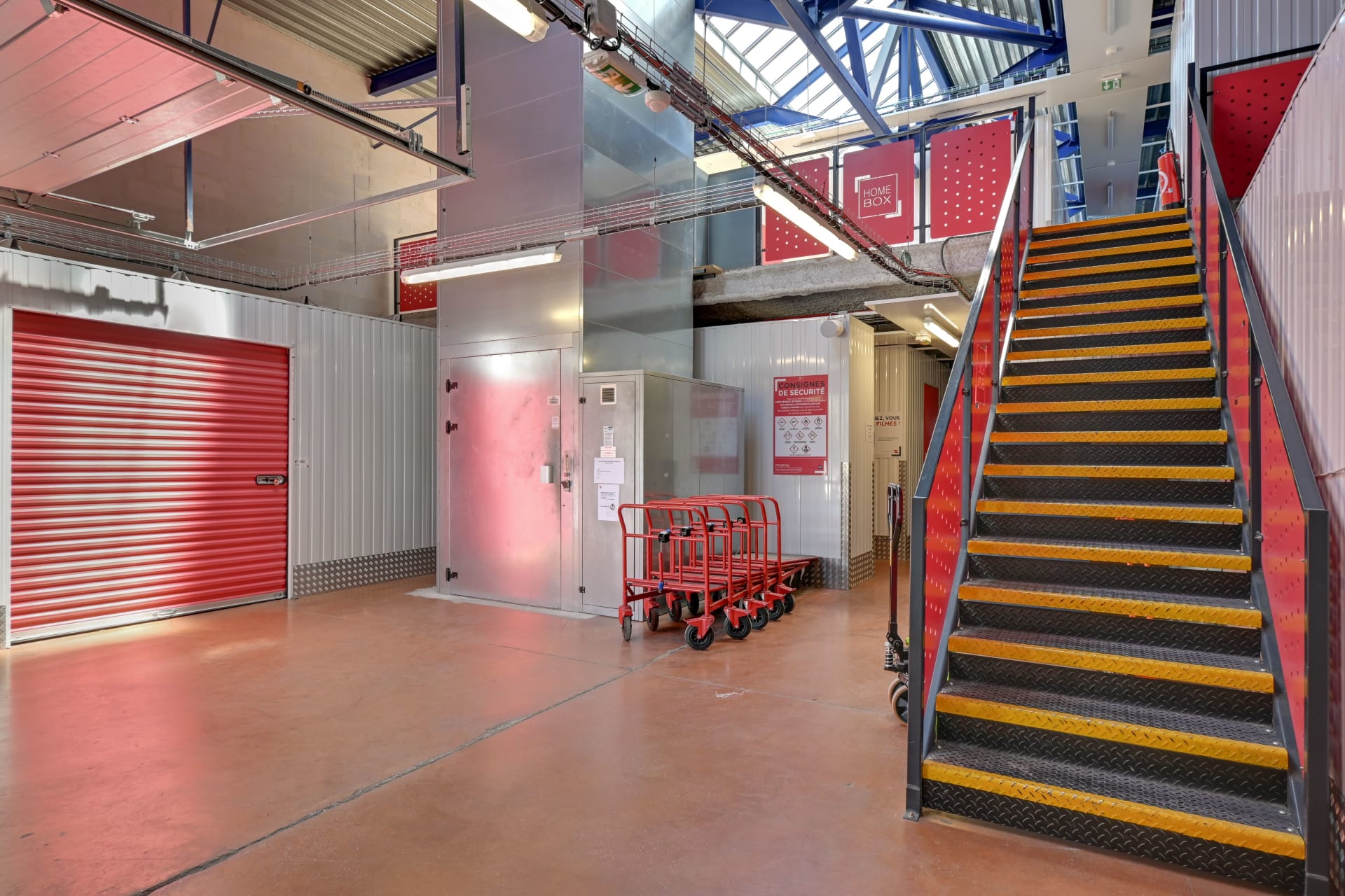 Couloir intérieur avec porte de box rouge, ascenseur vitré et escalier d'accès aux étages du centre HOMEBOX Toulon - La Farlède