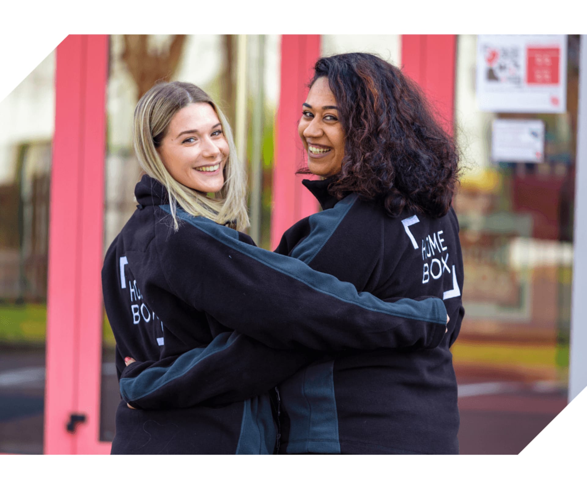 Deux collaboratrices HOMEBOX en sweat noir s'embrassant chaleureusement à l'entrée d'un centre de self-stockage