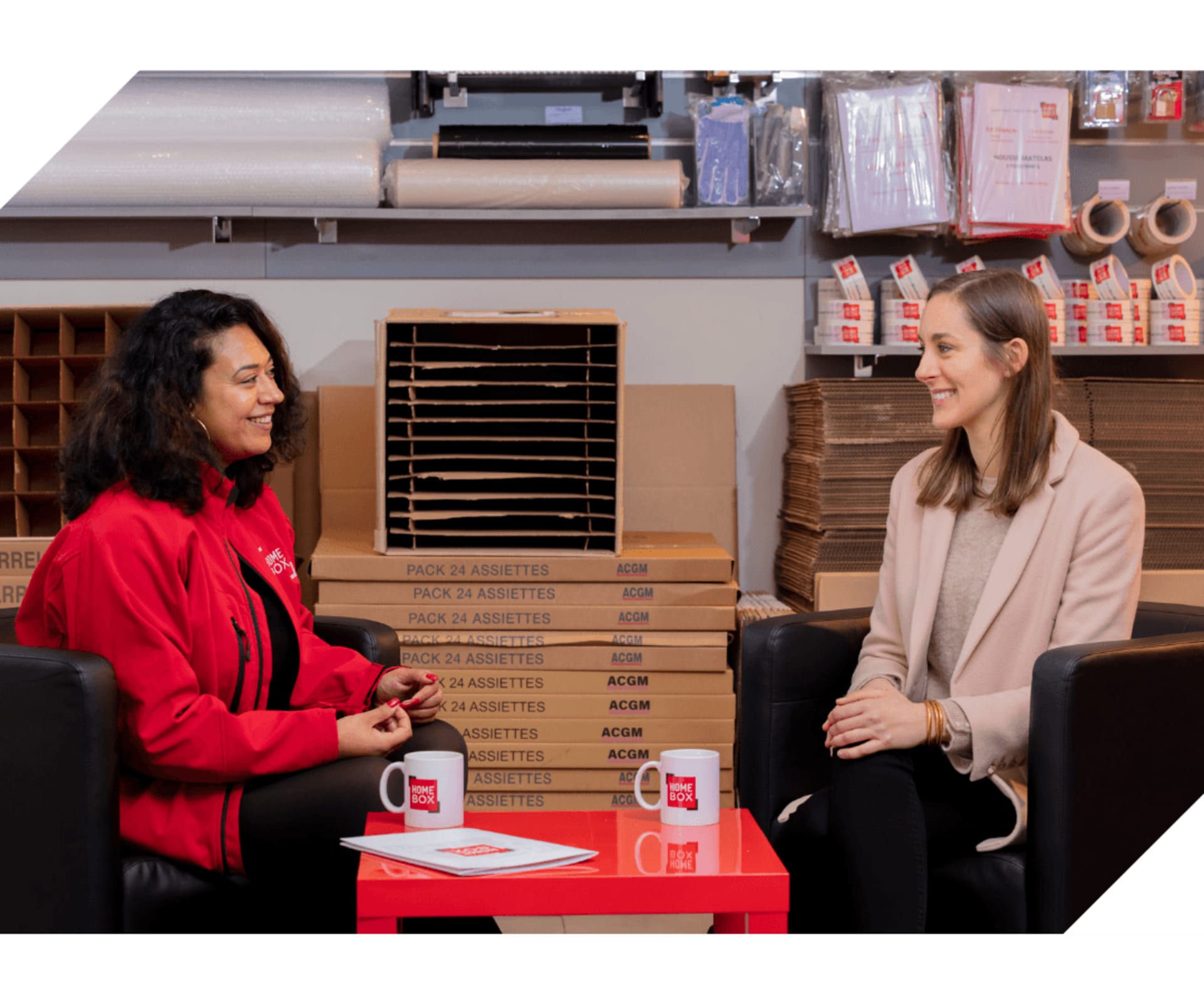 Deux femmes échangeant assises autour d'une table basse rouge dans l'accueil chaleureux d'un centre HOMEBOX