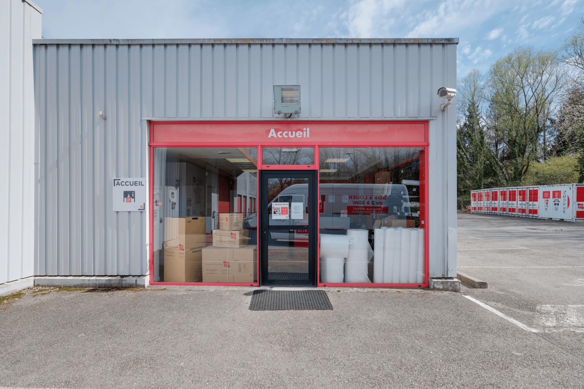 Façade et entrée de l’accueil du centre HOMEBOX Annecy, zone des Vernes à Cran-Gevrier, avec cartons et fournitures visibles à l’intérieur.