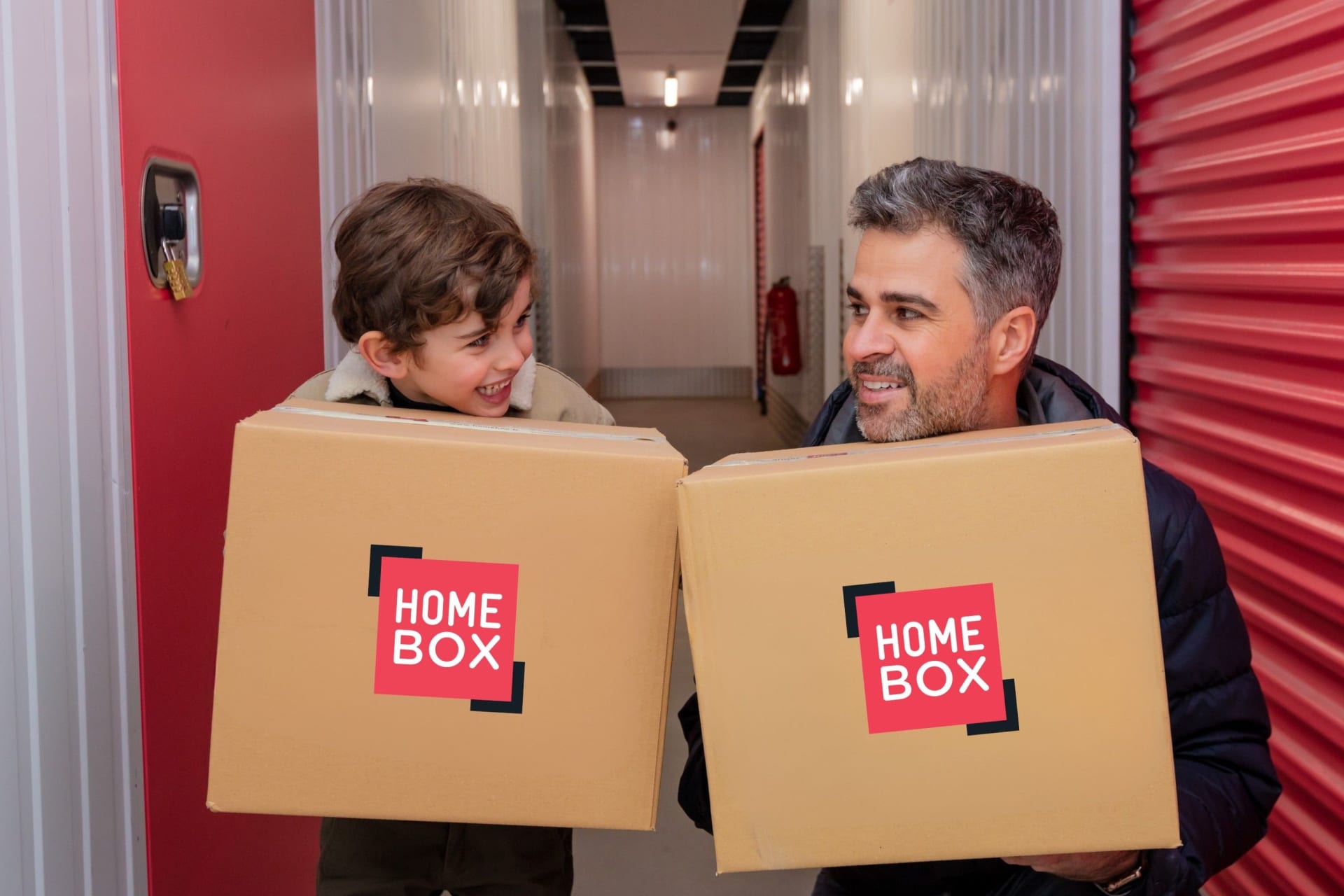 Un enfant et son père portant chacun un gros carton dans le couloir d’un centre de stockage. Ils se regardent, souriants.