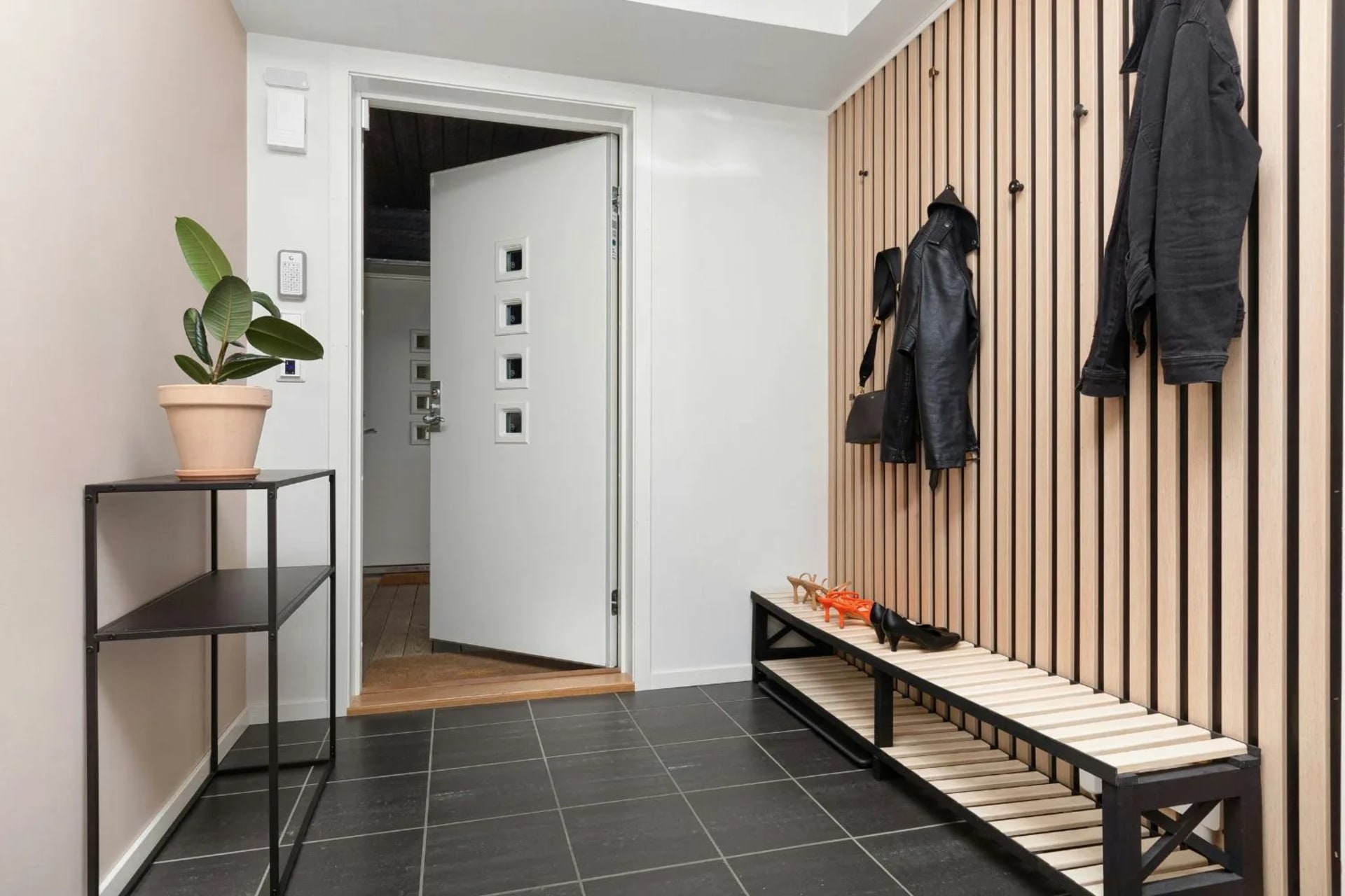 Hall d’entrée avec carrelage noir au sol, console industrielle noire à gauche, banc bois et métal noir à droite, claustra et patères noires à droite