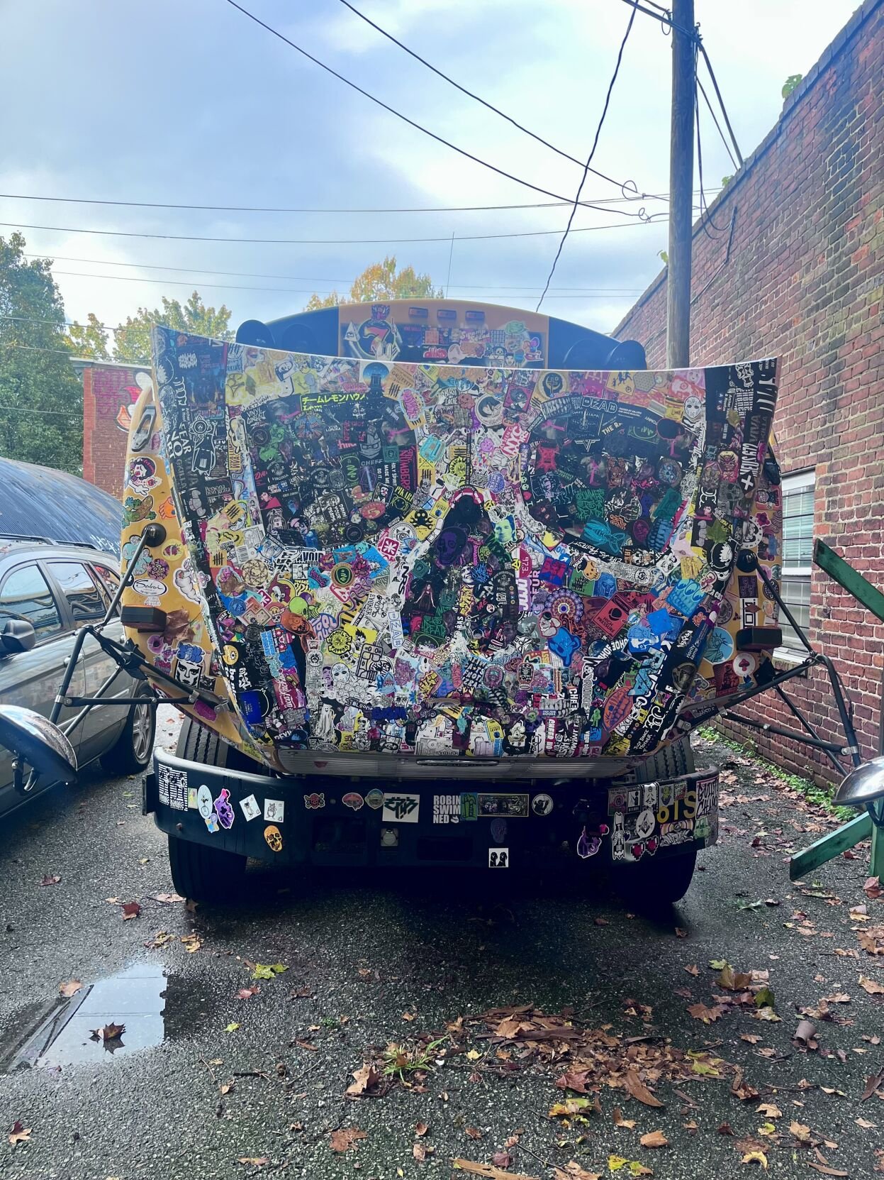 Front view of the 'Sticker Bus' with its hood completely covered in a sticker mosaic forming a large skull shape.