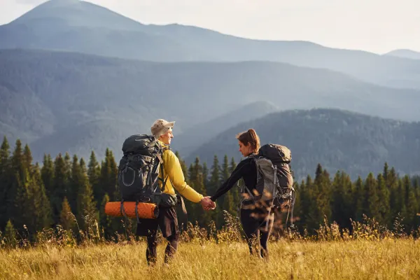 Two individuals with backpacks stroll through a lush green field under a clear blue sky.