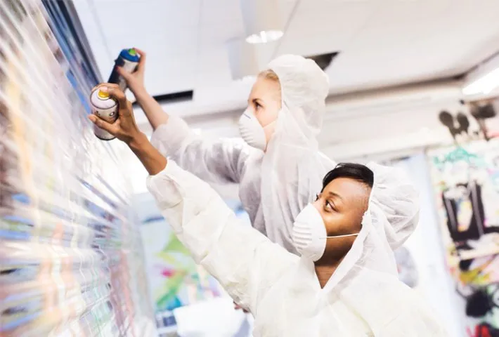 Graffiti artists at work: Two people in white protective suits and face masks spray painting a wall.