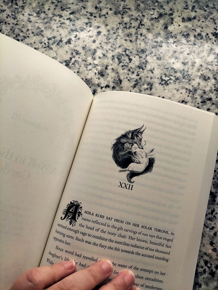 Photograph of a page inside the book. It shows the beginning of a chapter (XXII, the twenty second one), preceded by a small, greyscale drawing of a rabbit in a wolf's embrace, with the wolf holding a rose in its mouth.  The first paragraph it's completely visible, with the first letter adorned with flowers. It says, "AMIRA ROSE SAT HIGH ON HER SOLAR THRONE, its name reflected in the gilt carvings of sun rays that ringed the head of the ivory chair. Her brown, beautiful face emitted enough rage to outshine the merciless radiance of ten thousand burning suns. Such was the fury she felt towards the accused standing opposite her."  The second paragraph is partially visible, interrupted by my fingers. "Since word had travelled [...] the water of the attempt on her daughter's life she had [...] for [...] extradition. While [...] way of tenderness [...]."