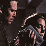 Wesley stands behind Faith, pressing a cold compress to the wounds and bruises in her face.