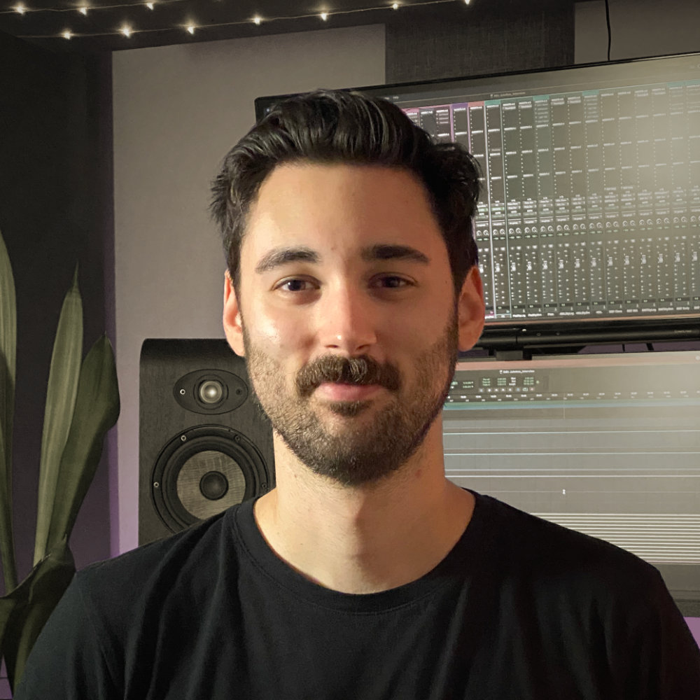 Luke Fair showing a friendly smile while sitting in his studio in front of studio monitors, screens showing Pro Tools, and a house plant.
