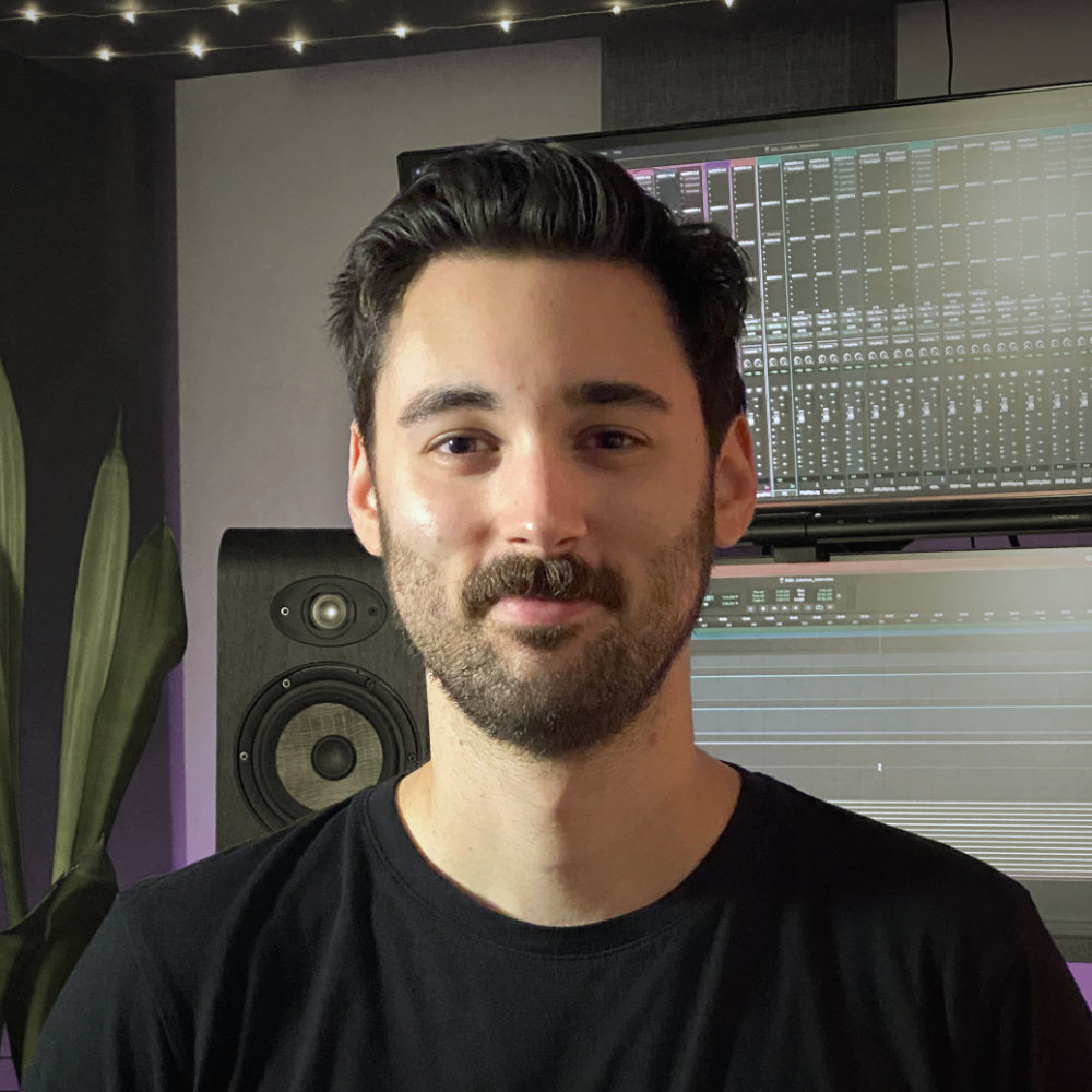 Luke Fair showing a friendly smile while sitting in his studio in front of studio monitors, screens showing Pro Tools, and a house plant.