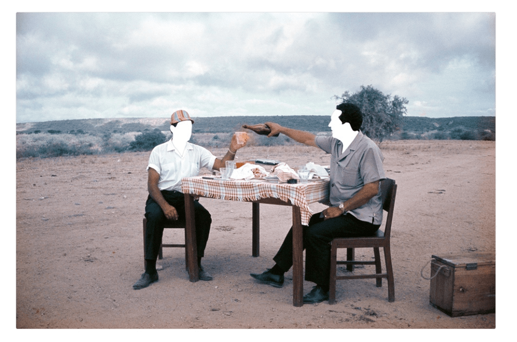 Two men at a table in a desert, faces blanked out.