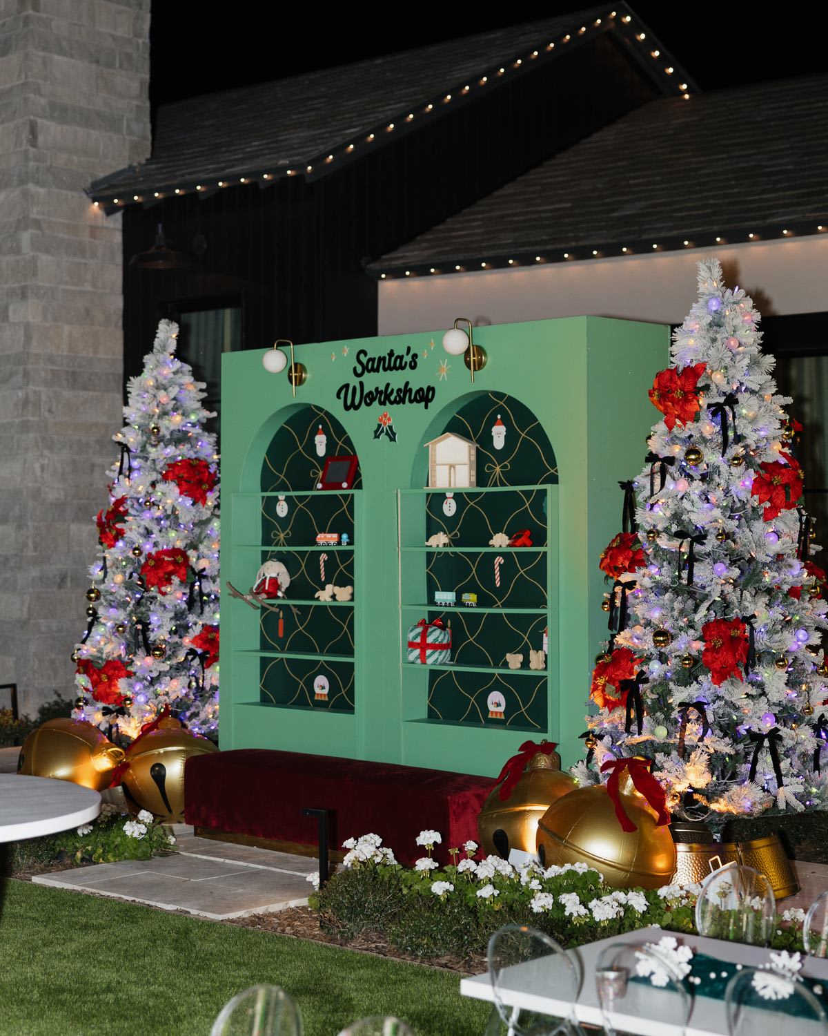 Frosted Christmas trees with red poinsettias beside a green workshop backdrop and twinkle lights on a backyard at dusk