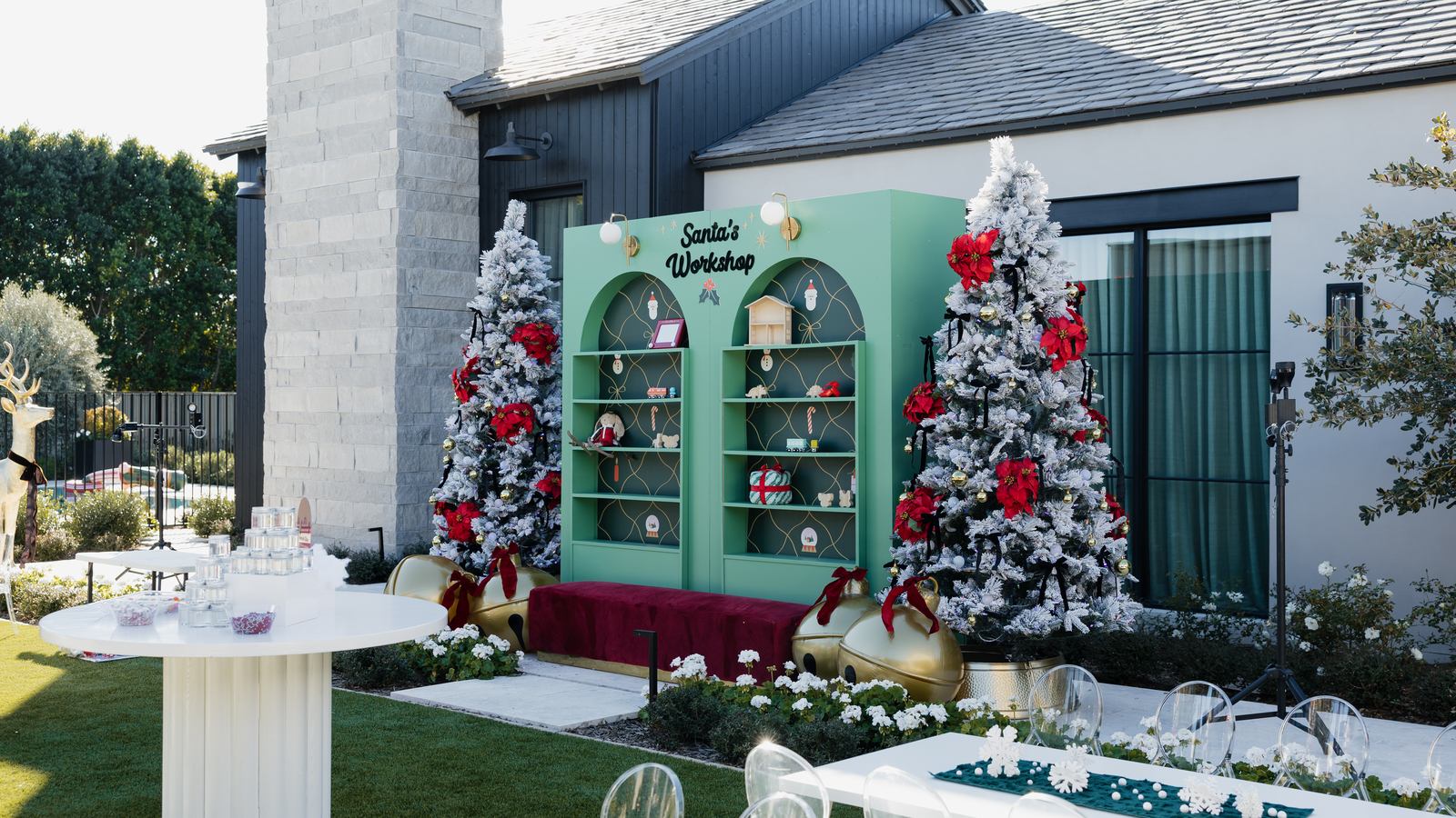 Backyard holiday party backdrop with green arched shelving styled as Santa's workshop, a red velvet bench and frosted Christmas trees with red poinsettias