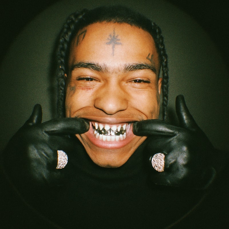 XXXTentacion. he haves face tattoos, tear tatoo. he is smiling at the camera wit