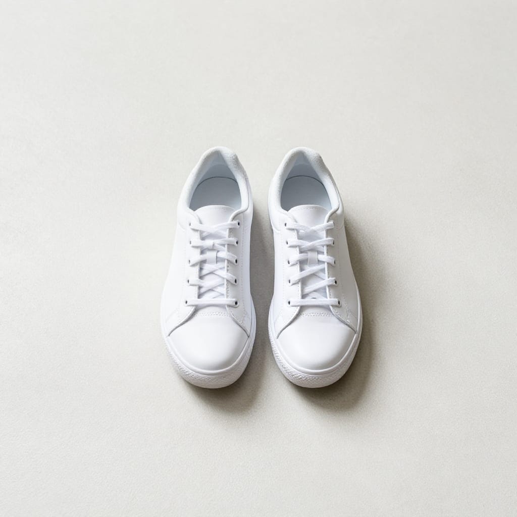 Overhead shot of pristine white sneakers resting on a smooth, light-colored concrete surface, captured with a wide-angle lens to emphasize their form and texture. The composition centers the shoes as the dominant subject, arranged in a precise line across the frame. Soft, diffused daylight from above creates gentle shadows beneath the soles, highlighting subtle details like stitching, laces, and reflective accents. The concrete background exhibits a faint, uniform texture with minor imperfections, enhanced by a slight sheen under the light. A quiet atmosphere prevails, with minimal movement or distractions, evoking a sense of calm precision and clean design. The color palette is minimalist, dominated by crisp white sneakers against a neutral gray concrete base, creating a serene, focused visual statement.
