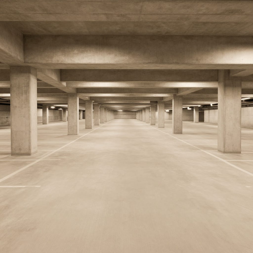 A wide shot of an expansive underground parking garage viewed from a high vantage point, capturing the vastness of the space. The scene is composed of evenly spaced concrete pillars arranged vertically, their straight lines converging toward a distant vanishing point in the background. A smooth, uniformly lighted concrete floor stretches beneath them, marked with clear grid lines for parking spaces. The lighting is artificial and consistent, emanating from rows of overhead fixtures that cast a soft, even glow across the ceiling and floor, eliminating shadows and creating a neutral, clinical atmosphere. The monochromatic palette emphasizes the stark, utilitarian texture of concrete, with no warm tones or highlights to soften the industrial feel. The composition is symmetrical and grounded, with no visible furniture or vehicles, reinforcing a sense of quiet stillness and isolation. The overall feeling is relaxed and contemplative, evoking a moment of quiet solitude in a space designed for function rather than life. The lens type is likely a standard wide-angle, capturing the full depth of the garage without distortion or compression. No motion is present, enhancing the stillness of the scene.