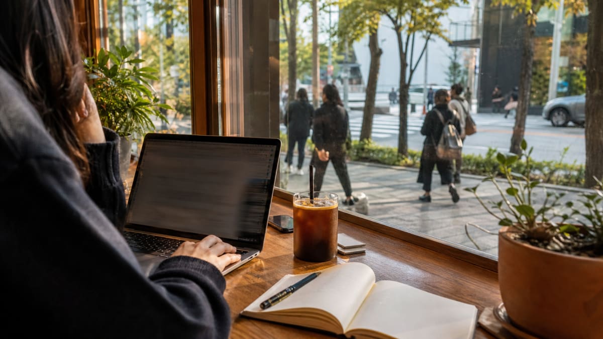 Person working on a laptop with an iced coffee beside a cafe window