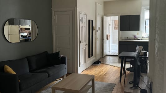 This cozy apartment features a compact and open layout. On one side of the room, there's a comfortable dark grey sofa with a yellow cushion, positioned against a darker grey accent wall. Above the sofa, a modern, oval mirror adds a touch of style and reflects natural light. In front of the sofa, a light wooden coffee table rests on a grey area rug, creating a warm and inviting atmosphere. 

The adjacent wall leads to a small kitchen area with white cabinetry and a sleek black countertop. A wooden dining table with two chairs is situated in between the living space and the kitchen, perfect for enjoying a meal or working from home. Beyond the dining area, a doorway leads to what appears to be a well-lit bathroom, complete with a tiled floor. The arrangement of furniture and use of neutral tones give the apartment a clean and modern feel, ideal for a comfortable stay.