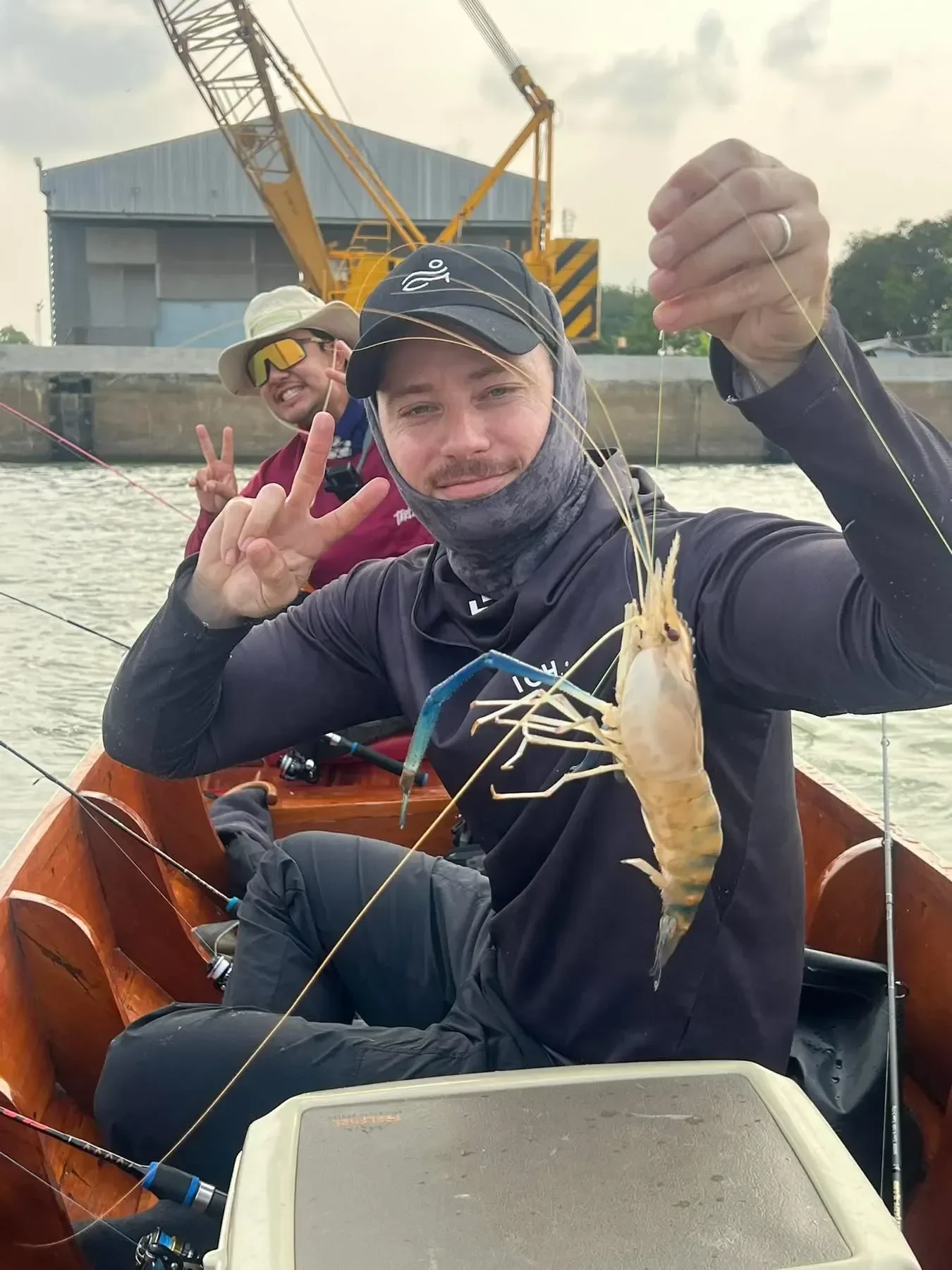 Angler smiling and holding a pale giant freshwater prawn while another angler sits behind him in a wooden boat.