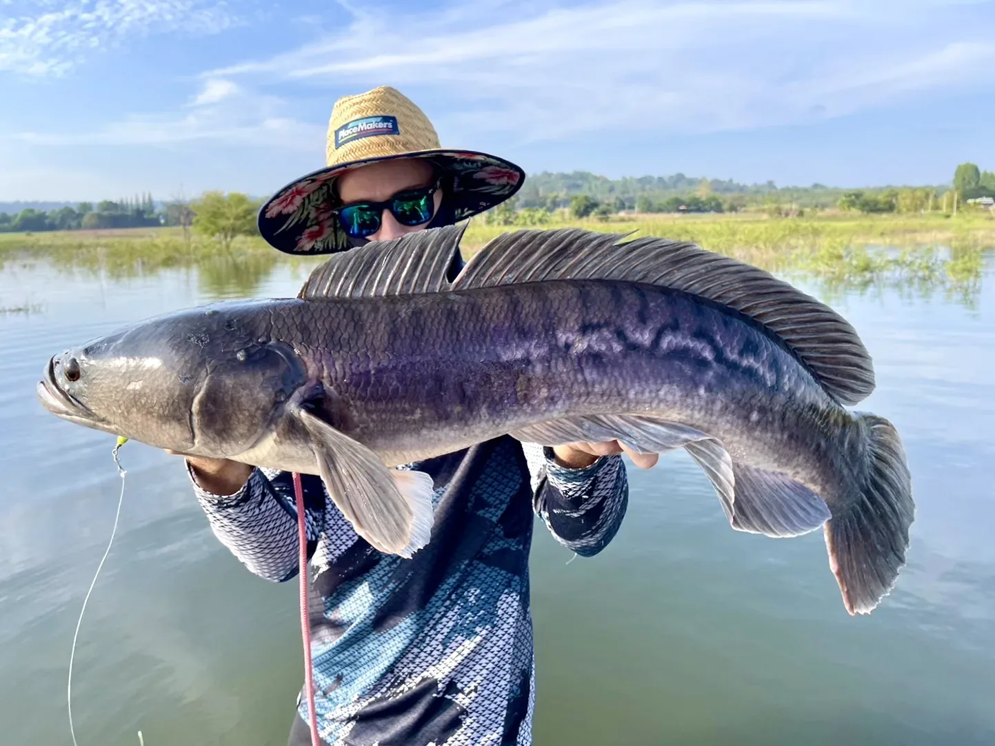 Angler in a straw sun hat and polarized sunglasses holding a large Giant Snakehead fish with both hands, standing in front of a calm lake in Thailand.