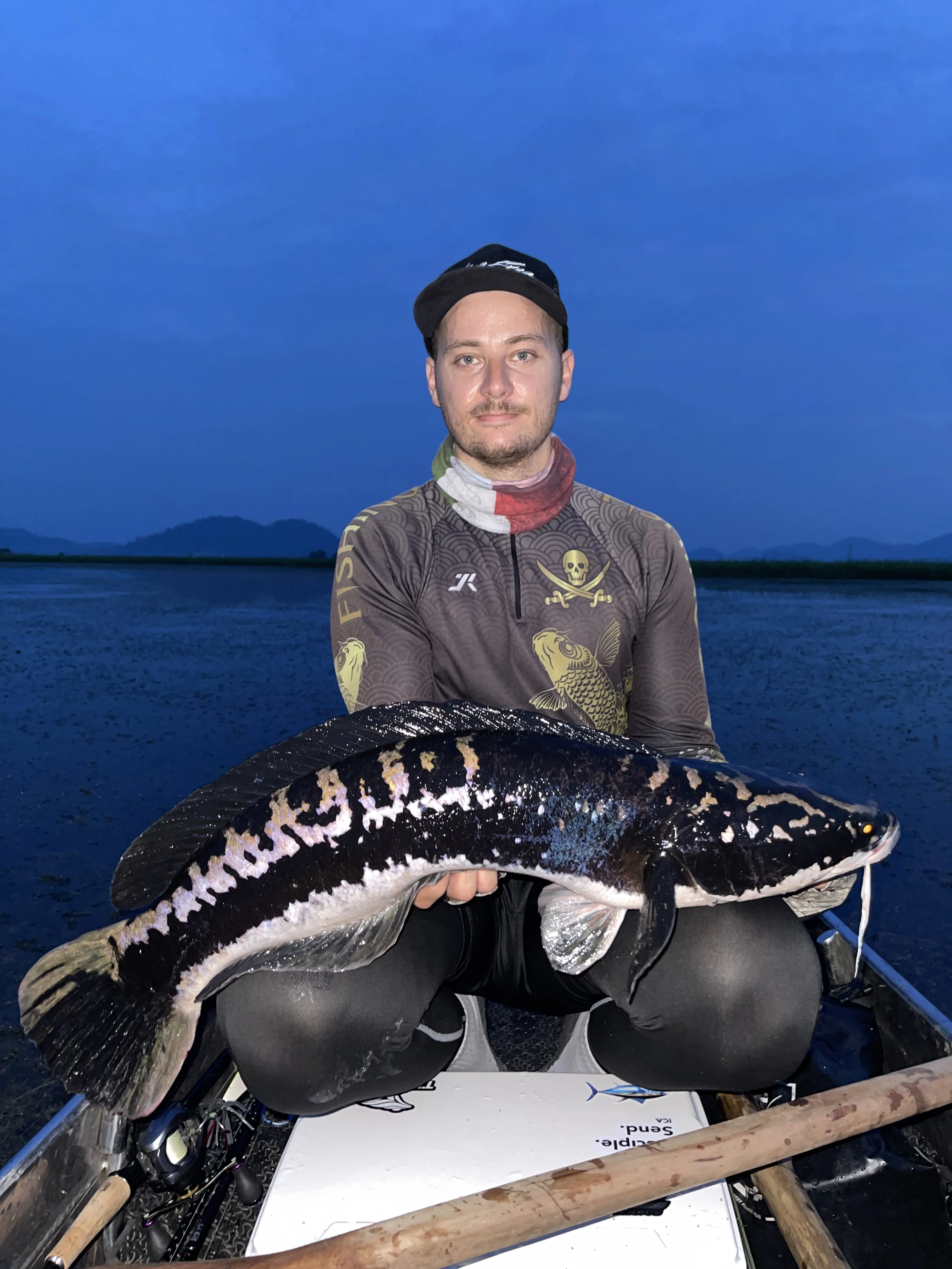 Angler seated in a small boat at dusk holding a large black-and-white patterned Giant Snakehead fish with mountains and dark blue sky in the background.