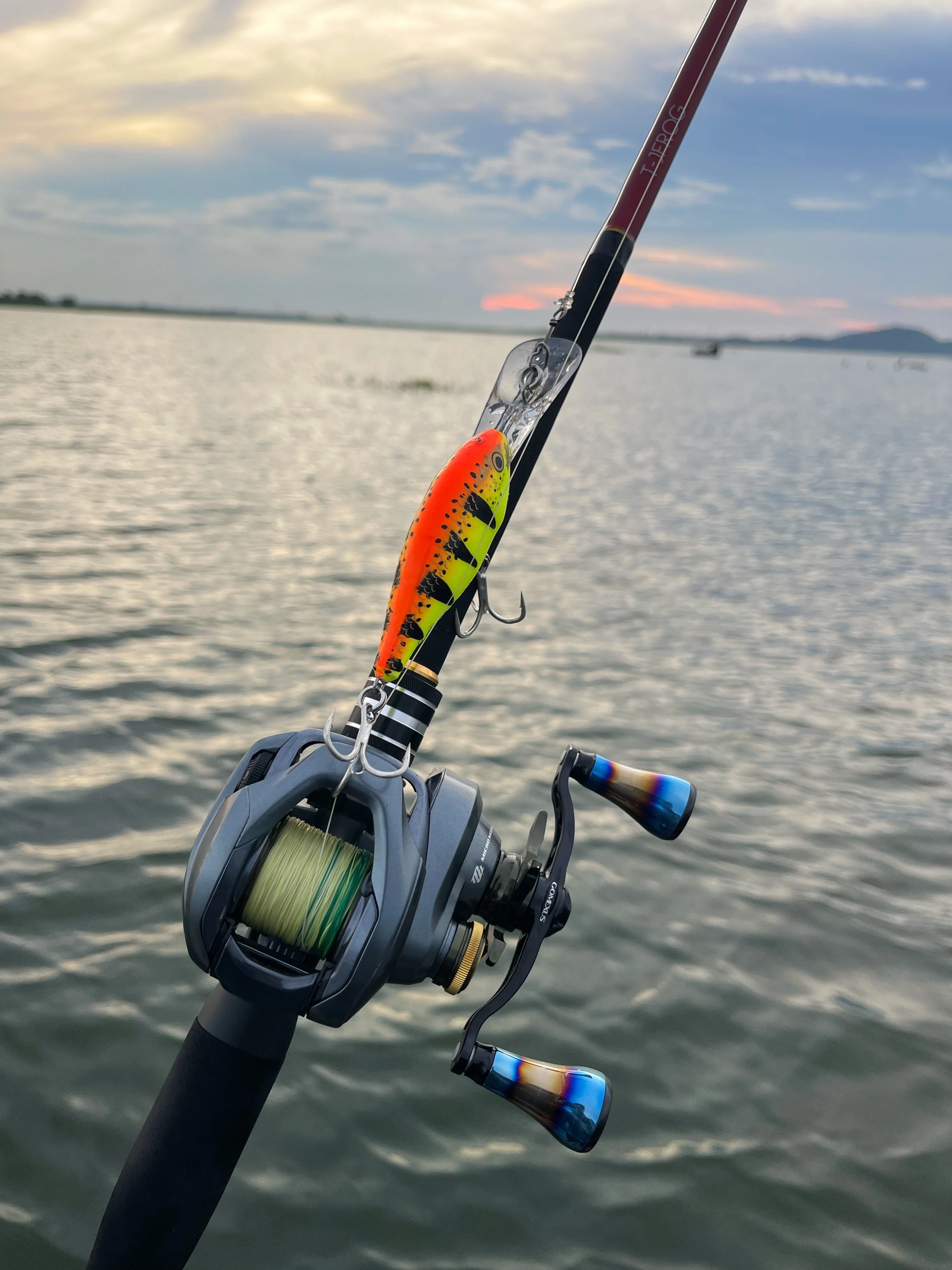 Close-up of a baitcasting rod and reel spooled with braided line, with a bright orange and yellow crankbait lure attached, against a colourful sunset over open water in Thailand.