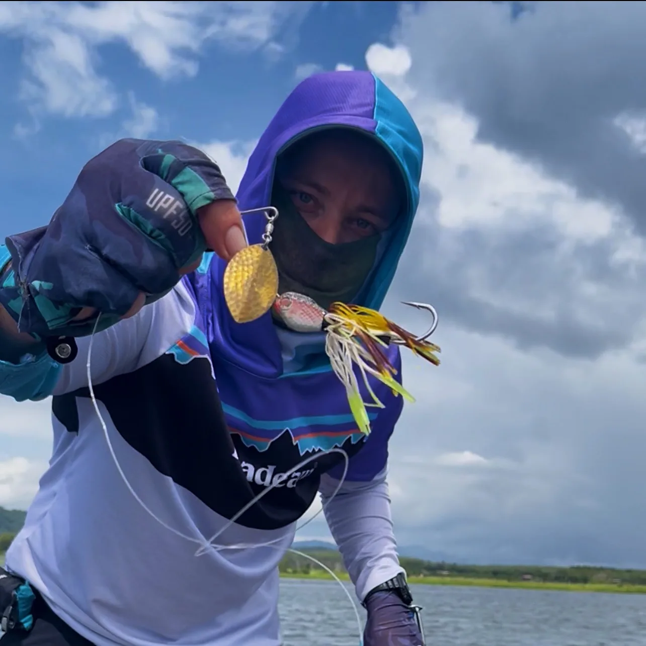 Close-up of an angler in a purple sun hood and UPF gloves holding up a gold spinnerbait lure with treble hook, on a boat over open water in Thailand.