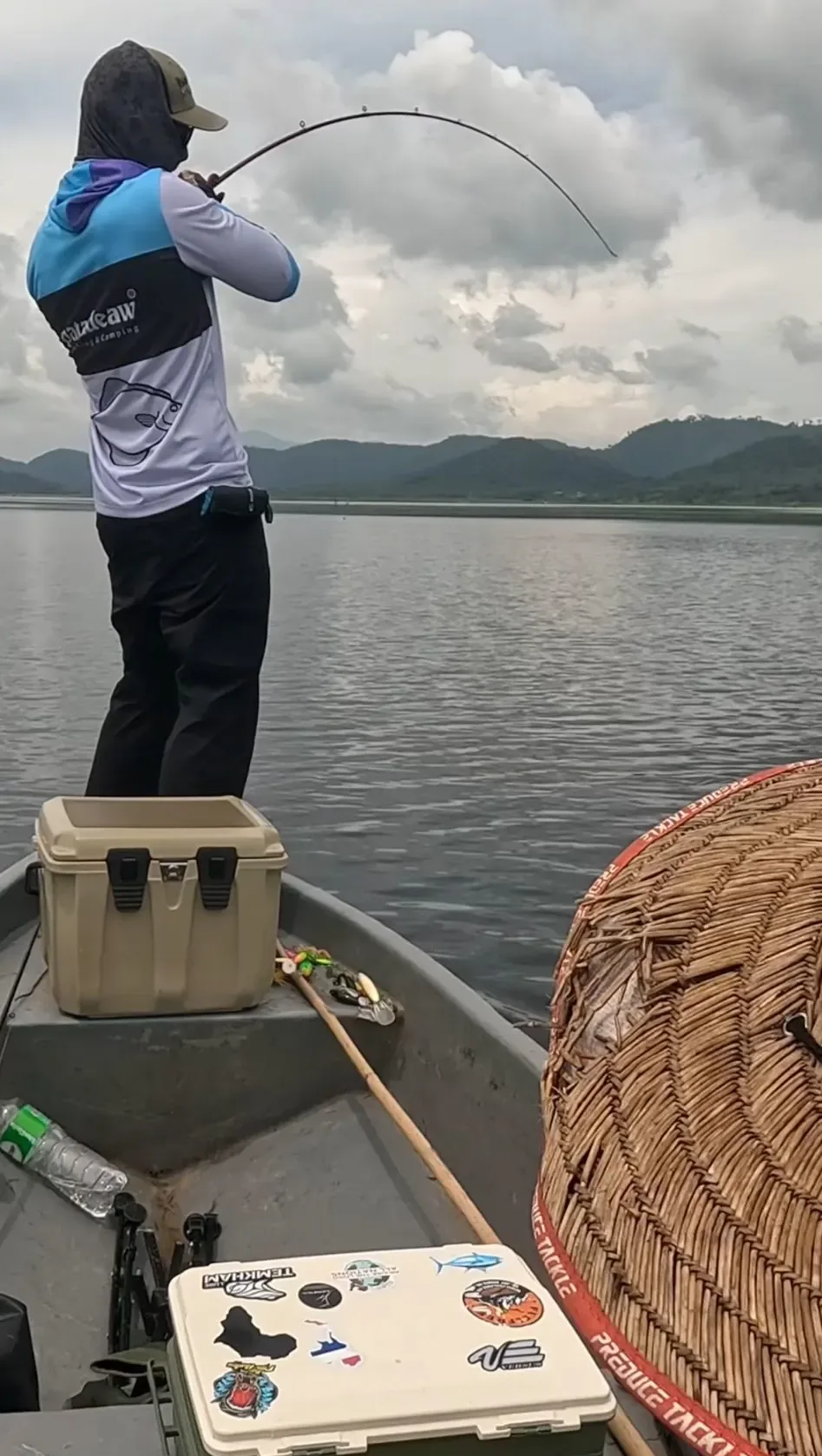 Angler standing in a boat on a Chanthaburi reservoir fighting a giant snakehead on a bent rod, with mountains and cloudy skies in the background.